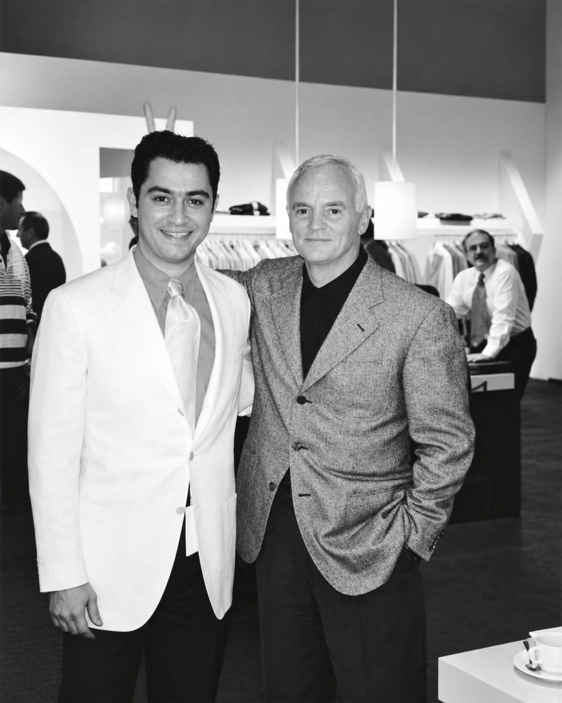 A black and white photograph of two men standing together in a clothing store. The man on the left wears a white blazer and light tie, and the man on the right wears a textured gray blazer over a black shirt.